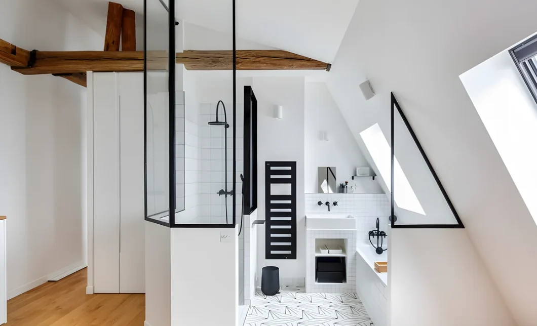 Salle de bain sous les combles avec douche fermée par une verrière noire, poutres apparentes et carrelage graphique