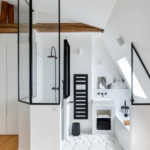 Salle de bain sous les combles avec douche fermée par une verrière noire, poutres apparentes et carrelage graphique
