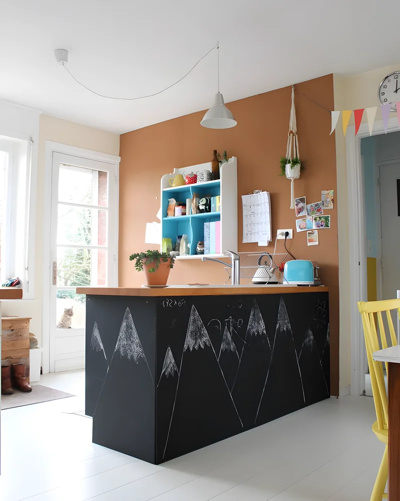 Cuisine lumineuse avec mur en liège, îlot noir décoré à la craie et étagères bleues