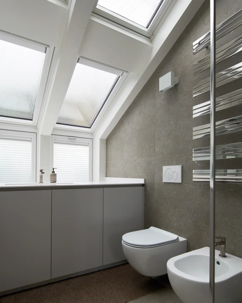 Toilettes modernes sous pente avec fenêtres de toit, mur effet béton, WC suspendu et meuble gris