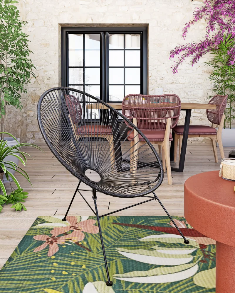 Fauteuil de jardin rond façon Acapulco en résine recyclée noire sur une terrasse fleurie avec tapis tropical